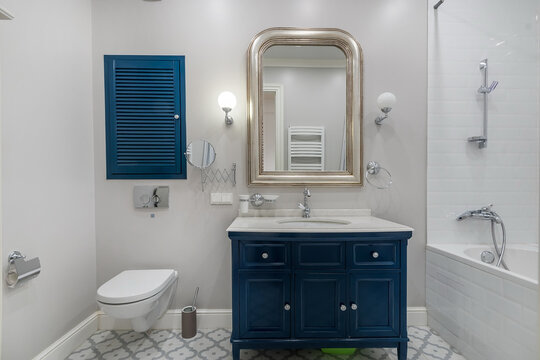 A well-designed bathroom shows a blue cabinet under a sink, a large mirror, and a shower area. Light from a wall fixture brightens the space. A toilet is also present along with a towel rack