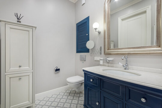 This bathroom features a blue cabinet, a white sink, and a large mirror. The walls are light gray and there is patterned flooring. Natural light enters the space