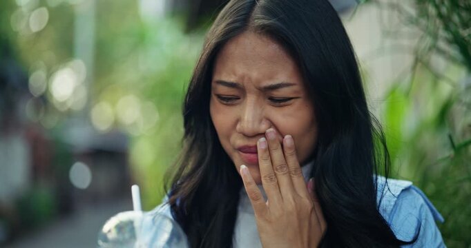 Young Asian woman experiences sudden toothache pain and holds her cheek, suffering from sensitive teeth after drinking a cold iced beverage outdoors in a sunny park.