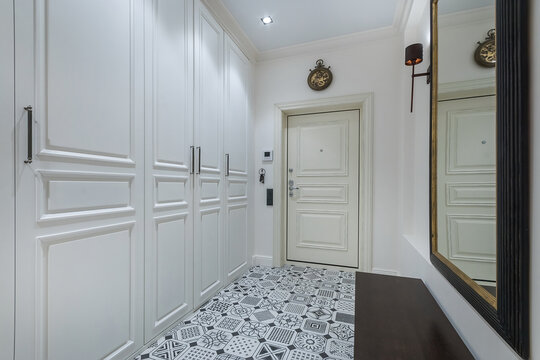 The hallway has white cabinets and a door leading outside. There is a mirror on the wall and patterned tiles on the floor. Wall decorations add a simple touch to the space
