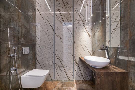A bathroom shows a sink on a wooden cabinet, a toilet, and a glass shower. The walls have marble tiles, and ceiling lights provide bright illumination