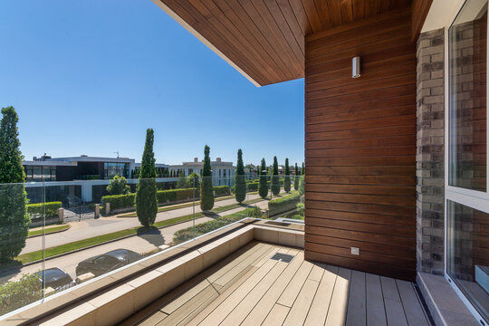 A balcony overlooks a street lined with trees and modern homes. The sun shines brightly, highlighting the clear blue sky and the greenery. Vehicles are parked along the street