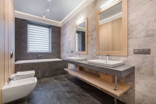 A bathroom features a tub, two sinks, and mirrors. Sunlight enters through a window. The walls have light stones, and wood accents are present. Clean lines and functional space are visible