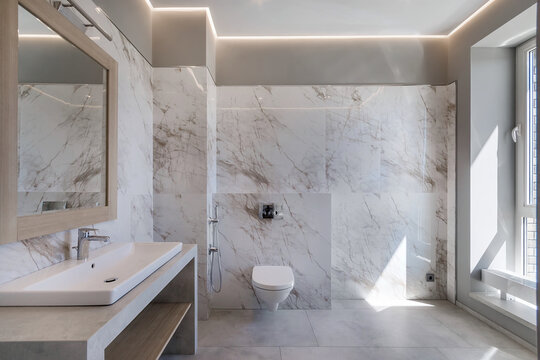This bathroom features marble walls, a large mirror, and a modern sink and toilet. Natural light fills the space, highlighting the clean lines and simple design