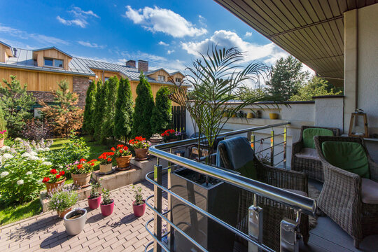 The balcony features green plants and colorful flowers in pots. Behind it, there are buildings and trees under a clear blue sky during midday