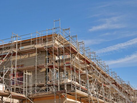 low-angle shot of a multi-story residential building completely enveloped in metal scaffolding during a major renovation or construction phase. The structure features wooden planks and safety rails un