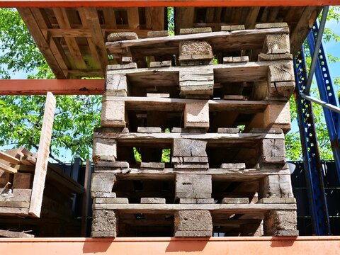 medium shot of weathered wooden pallets stacked on an orange metal industrial rack outdoors. The worn timber shows deep textures and age, set against a background of green trees and a clear blue sky. 