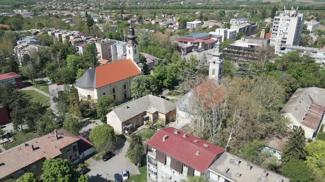 Cinematic aerial pull-back revealing a wide panorama of Vrbas, northern Serbia. Lush greenery and parks integrated into the urban cityscape under a bright sunny sky.