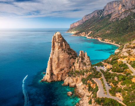 Aerial view of rock and mountains, blue sea bay, beach, boat on sunny summer day. Sardinia Island, Italy. Top drone view of cliffs, stones, green trees, road, transparent water at sunset. Landscape