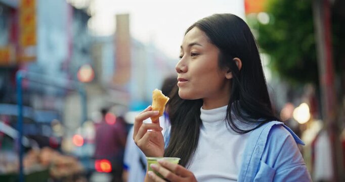 Happy young Asian woman with closed eyes enjoys eating a delicious deep fried dough street food snack with green dipping sauce on a busy urban city street outdoors.