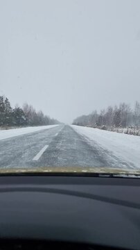 POV: driving on asphalt road during snowfall. Icy two-lane road during a blizzard: falling snow obscures the horizon and forest - hazardous driving conditions.