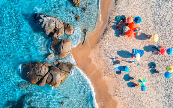 Aerial drone view of colorful umbrellas, sandy beach, blue sea with waves and rocks at sunset. Summer in Sardinia island, Italy, Tropical seascape. Top down view of clear water, relaxing people, beach