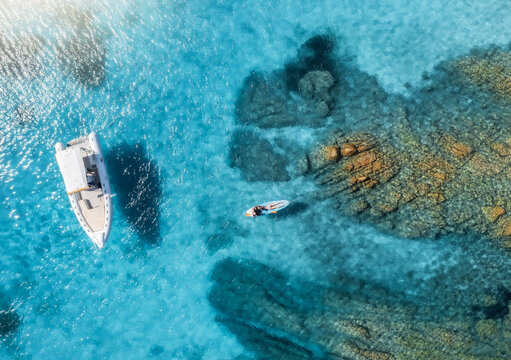 Aerial view of a woman practicing paddle boarding in beautiful turquoise waters next to a rocky coast and aboat, enjoying a sunny summer day. Top down drone view of sup board, boat and clear blue sea