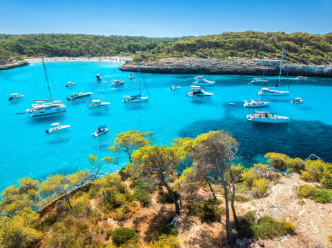 Aerial drone view of yachts and boats anchored in turquoise sea bay surrounded by rocky coastline and green trees, summer vacation scene in Cala Mondrago, Mallorca, Balearic Islands, Spain. Top view