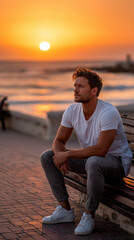 Fototapeta premium Young man with a charming smile looking at the camera, sitting casually on a bench on a waterfront promenade with the setting sun creating a warm golden glow over the ocean