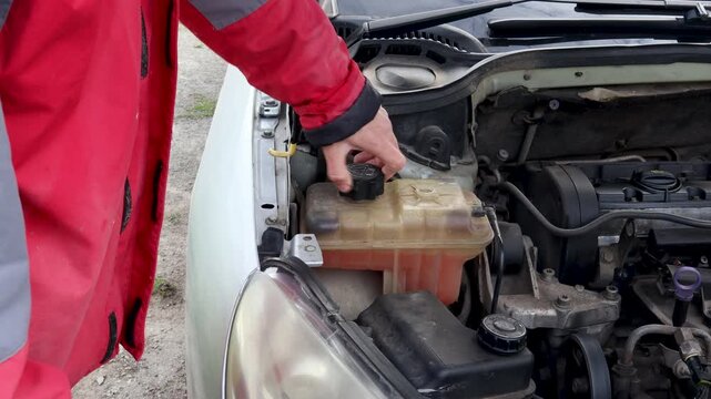 Mechanic opening the coolant reservoir tank on a car engine for a vehicle maintenance checkup, showing the radiator system and fluid level inside the expansion bottle