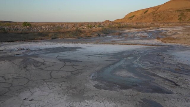 Arid landscape with dried cracked earth, mudflows, and a gentle hill in the background under clear sunset light. Natural environment of a mud volcano..