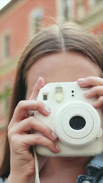 Vertical shot of Caucasian woman holding instant camera close to face. Female taking photo while covering eyes with device. City street background. Woman capturing moment during walk outside.