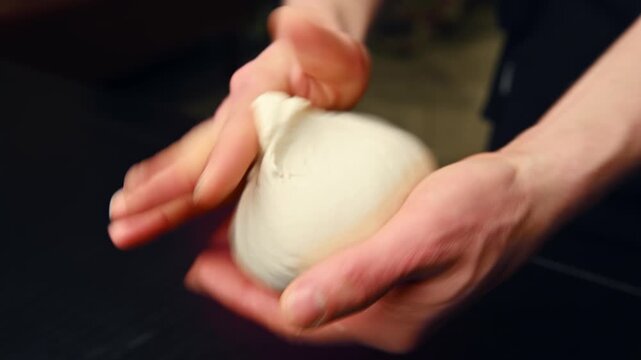 Hands kneading dough into a smooth ball on a dark countertop surface
