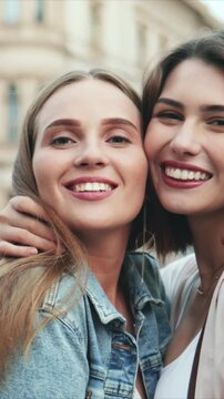 Vertical close view of two Caucasian women posing together. Female friends smiling at camera with cheerful mood. Street background. Women embracing and enjoying friendship while standing outside.