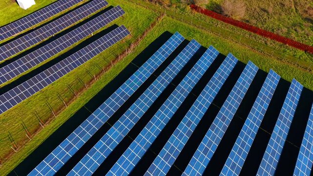 Solar power station with rows of panels on grass field. Wide aerial shot showing a solar panel array separated by a fence and green landscape under bright sun.