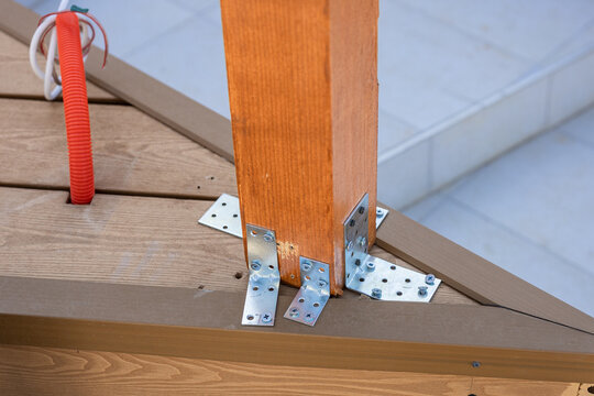 Close-up of a wooden beam connection reinforced with metal brackets and screws, showing structural detail of a roof or terrace construction. Electrical conduit visible on timber decking.