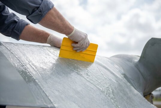 Worker hands using a yellow plastic squeegee to spread wet resin over fiberglass cloth material on a curved mold