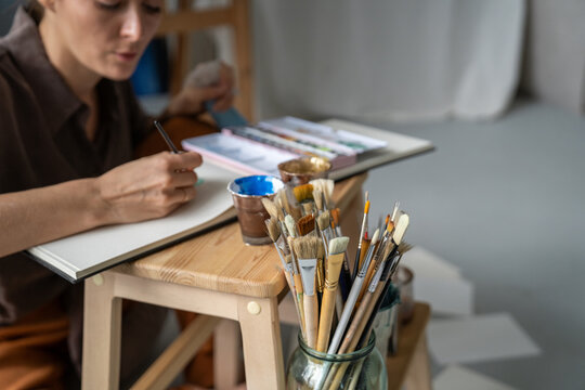 Serious focused woman painter drawing pictures at home using watercolors and sketchbook, female artist fully engaged in creative process, creating sketches, getting inspiration for future paintings.