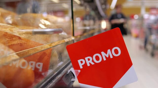 Close up of a red promotional sign in a supermarket bakery section, with fresh bread on the shelves and blurred customers walking in the background aisle, showcasing grocery store sales