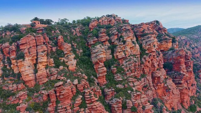 Red sandstone rock formations with small hermitage on mountain summit. Close view of stratified red sandstone pillars with dense green vegetation and a small stone building on top.