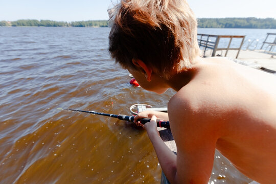 Boy leaning over wooden dock fishing with rod and holding small cup. Curious child watches water intently on sunny summer day at lake. Active youngster discovers excitement of angling outdoors