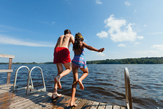 Father and daughter jump off a wooden dock into a lake on a sunny summer day. Man in red trunks with girl in blue swimsuit leaping together. Family shares thrilling outdoor recreation and bonding