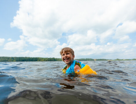 Boy swims in a lake wearing a bright blue and yellow life jacket on a sunny summer day. Child with a big smile enjoying the cool water. Kid experiences joyful outdoor recreation and water safety