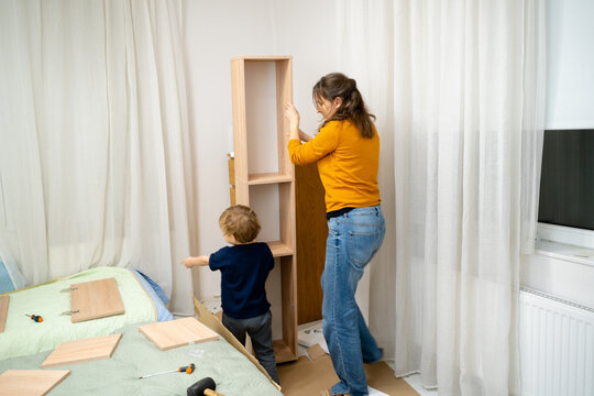A mother and her young son work together to position a newly assembled shelving unit in its place. Teamwork during the final stage of a DIY furniture project at home.