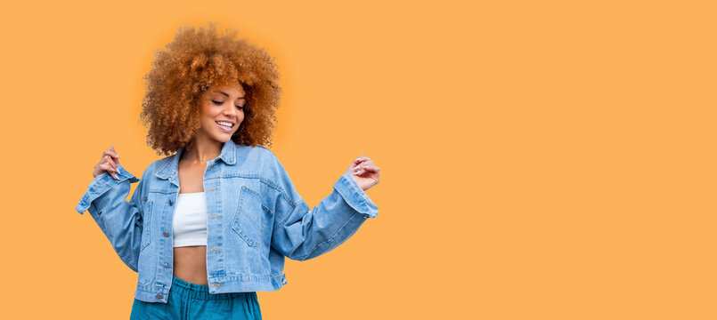 Playful happy young African American woman wear stylish denim clothes dancing and jumping funky black teen fashion girl on orange background. Curly hair student having fun. Web banner, copy space