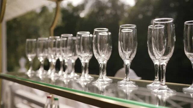 Row of clean, empty champagne glasses on glass shelf of gold bar cart. Ready for elegant party or wedding reception. Natural light, outdoor setting with green foliage background. Celebratory mood