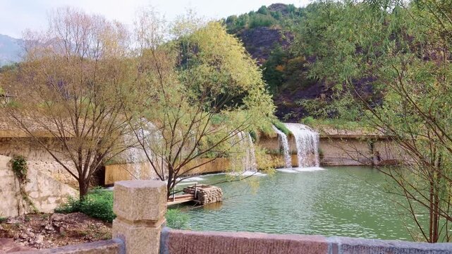 Landscape documents calm green water surface with small dam willow branches rocky hillside and forested slopes under clear daylight sky, Gubei Water Town, China, 4k