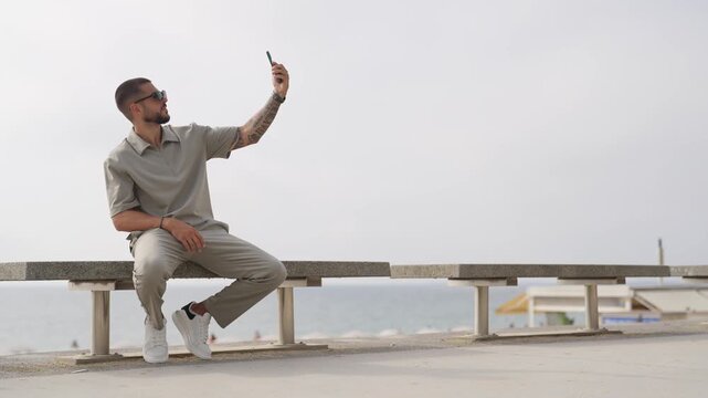 Man smiles while capturing selfie on bench. He wears casual outfit and sunglasses. Ocean background hints relaxed seaside vibe. Ideal for social media or lifestyle content