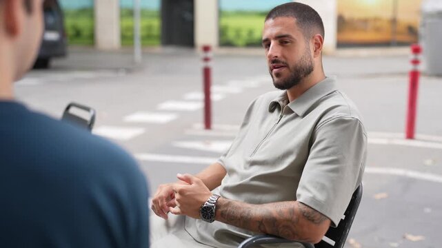 Man gestures while seated outdoors. Another person faces him, partially visible. Background shows parked cars and red bollards. Scene suggests casual conversation or interview