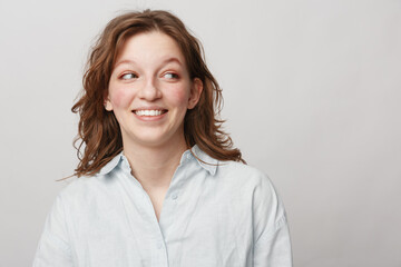 Fototapeta premium Smiling young woman with curly hair wearing white shirt looks sideways with happy expression on light background. Concept of positive emotions, casual style, and natural beauty.