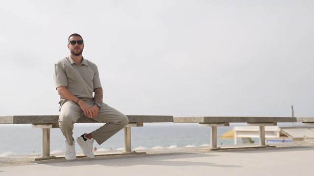 Man sits on bench, gazing at ocean. He wears casual clothes, sunglasses, and white sneakers. He looks relaxed, thoughtful, or perhaps slightly lost in thought