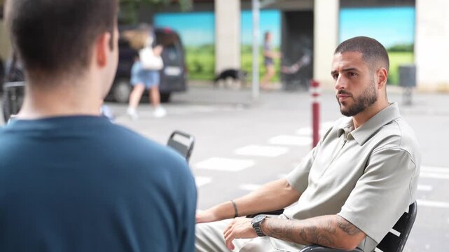 Man smiles warmly while seated in wheelchair outdoors. Conversation unfolds between two men on city street. Background shows pedestrians and parked vehicles