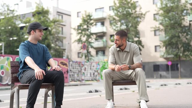 Two men sit on a bench in an urban park. They converse casually, showing relaxed expressions. Trees and buildings form the background. Perfect for social, urban, or lifestyle projects