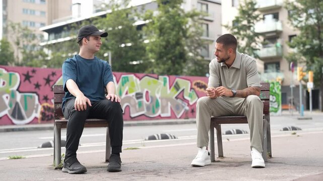 Two men chat on a city bench. One adjusts his cap while the other listens. Graffiti adds urban color to their setting. They seem relaxed, engaged in casual conversation