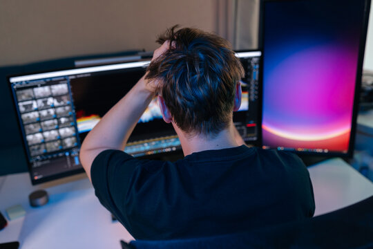 Man feeling stressed and frustrated, gripping head while working on computer with multiple screens in dark home office, experiencing burnout and overwhelming deadlines late at night.
