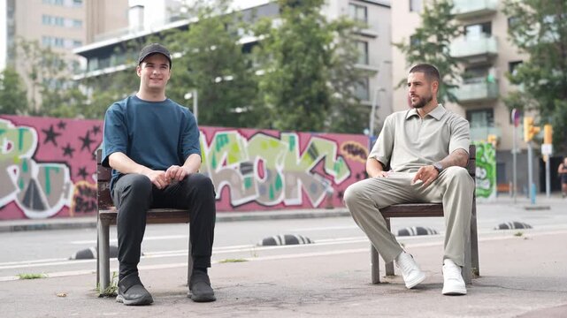 Two men converse on city benches. One smiles, the other gestures thoughtfully. Graffiti lines the wall behind them. Urban backdrop with distant buildings