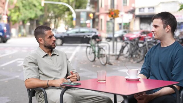 Two men converse at a cafe table. They sit facing each other, relaxed and engaged. City life buzzes softly behind them. Glass of water and a teacup sit quietly on the red table