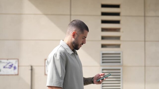 Man walks past a wall, focused on his phone. He glances to the side, showing awareness of surroundings. Soft light highlights his calm, concentrated expression. Urban scene, casual modern lifestyle