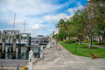 Obraz premium Miami, USA, 16 April 2026: Paved walkway along Miami marina with docked motor boats. Wide view of a concrete pier and green lawn with palm trees overlooking a boat marina in Miami, Florida, USA.