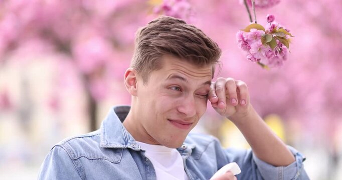 Seasonal allergy. Man with paper tissue sneezing near blossoming tree on spring day